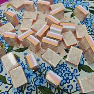 May include: A collection of rectangular tiles featuring a layered design with a translucent, marbled beige base topped by horizontal stripes of white, light pink, and soft orange. Each tile has a smooth, glossy finish and uniform shape.