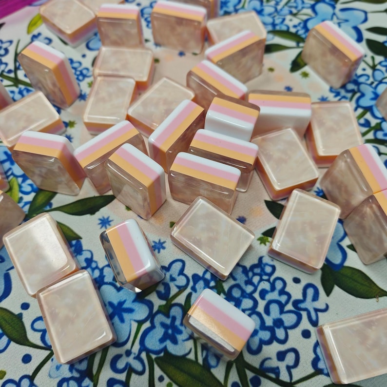 May include: A collection of rectangular tiles featuring a layered design with a translucent, marbled beige base topped by horizontal stripes of white, light pink, and soft orange. Each tile has a smooth, glossy finish and uniform shape.