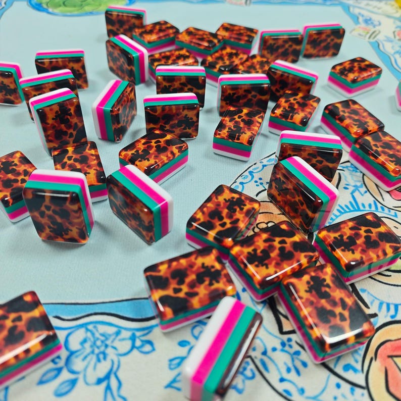 May include: A collection of rectangular mahjong tiles featuring a glossy brown and black tortoiseshell pattern on the top surface, with layered sides in vibrant shades of hot pink, teal, and white, scattered across a light blue patterned surface.