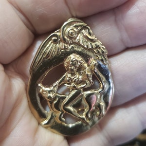 May include: Gold-toned pendant featuring a detailed relief of a winged figure riding a mythical creature. The pendant is oval-shaped and has a textured surface.