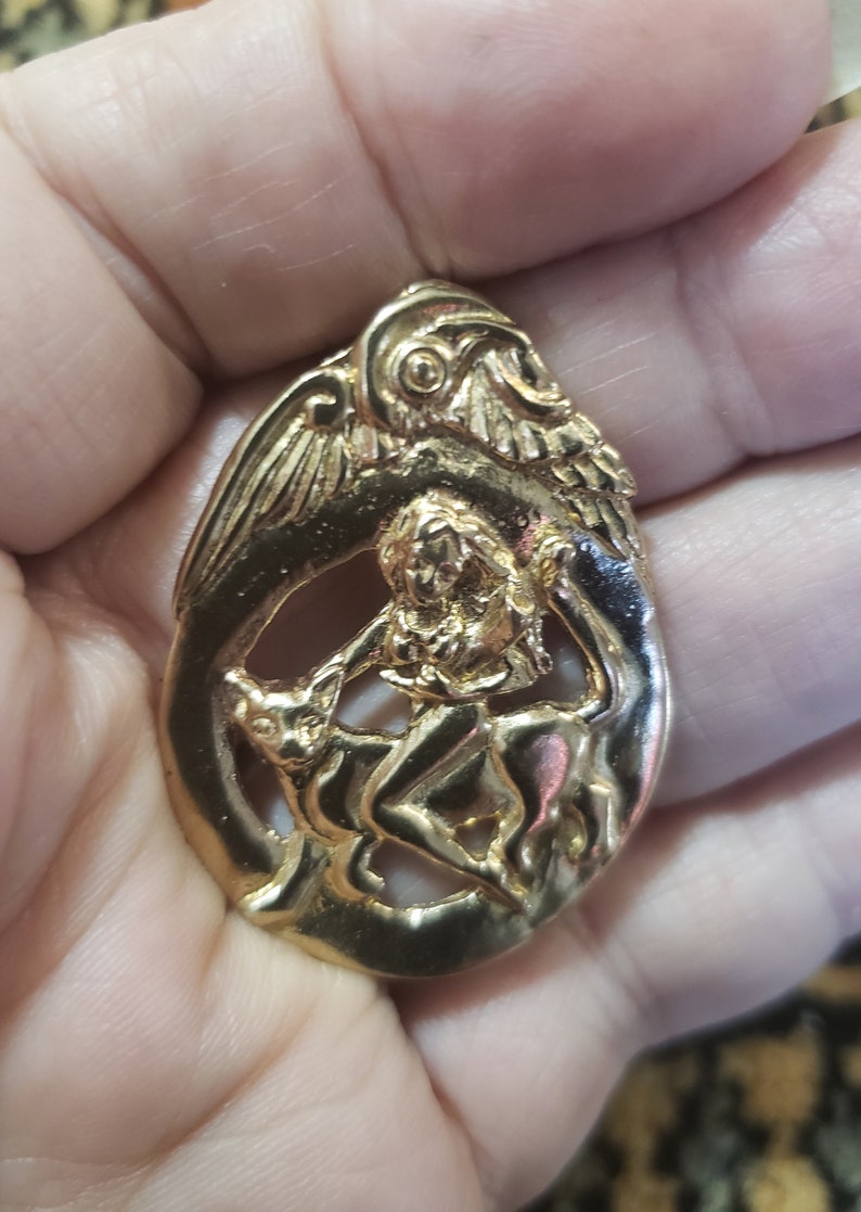 May include: Gold-toned pendant featuring a detailed relief of a winged figure riding a mythical creature. The pendant is oval-shaped and has a textured surface.