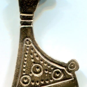 Puede incluir: Un colgante de hacha de color plateado con detalles intrincados. La cabeza del hacha está decorada con un patrón repetitivo de pequeños círculos y un círculo más grande en el centro. El mango del hacha es curvo y tiene un bucle en la parte superior para colgar.