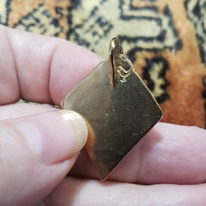 Puede incluir: Un colgante de color dorado en forma de diamante con un bucle en la parte superior para sujetar una cadena.