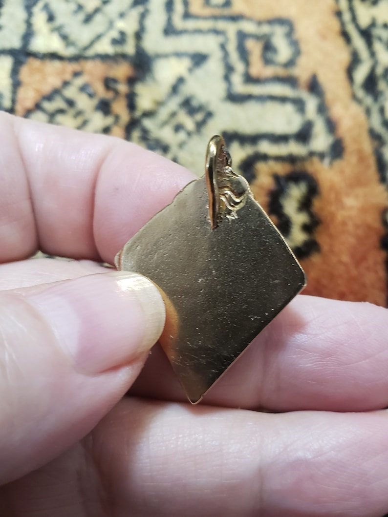 Puede incluir: Un colgante de color dorado en forma de diamante con un bucle en la parte superior para sujetar una cadena.