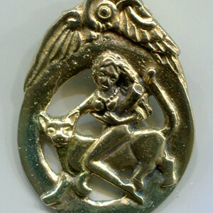 May include: A gold-colored metal pendant depicting a winged figure riding a cat. The figure is holding a staff and has a flowing robe. The pendant is oval-shaped and has a detailed design.
