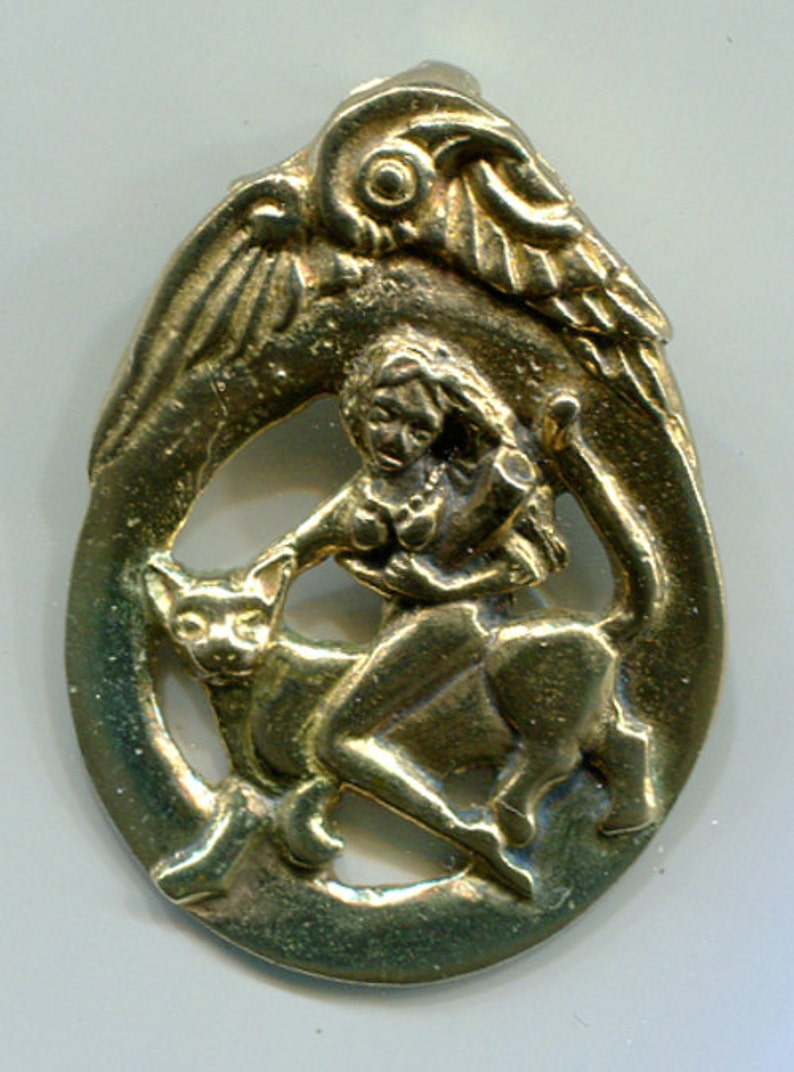 May include: A gold-colored metal pendant depicting a winged figure riding a cat. The figure is holding a staff and has a flowing robe. The pendant is oval-shaped and has a detailed design.
