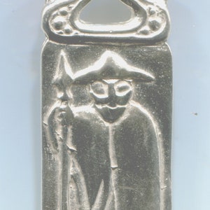 May include: A silver pendant with a detailed design of a figure holding a spear. The figure is wearing a hat and a long robe. The pendant has a loop at the top for hanging.