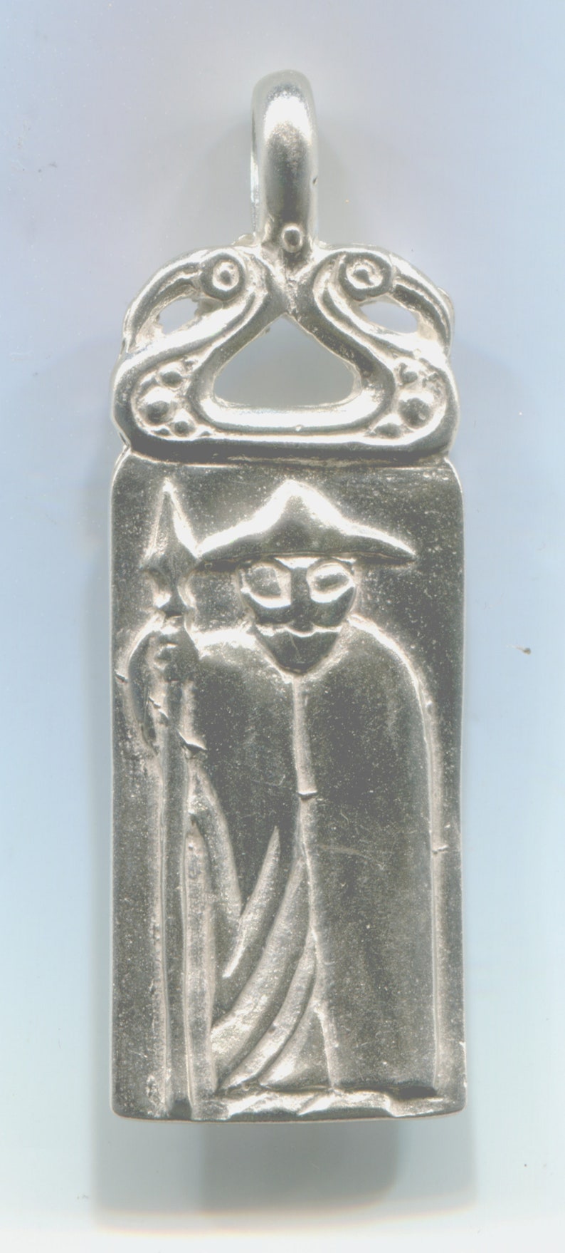 May include: A silver pendant with a detailed design of a figure holding a spear. The figure is wearing a hat and a long robe. The pendant has a loop at the top for hanging.