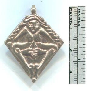 Puede incluir: Un colgante de color dorado en forma de diamante con una imagen en relieve de una figura femenina desnuda con el pelo largo. La figura est&aacute; sentada con las piernas cruzadas y las manos juntas delante de ella.