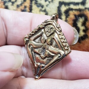 Puede incluir: Un colgante dorado con una escultura en relieve detallada de una figura femenina desnuda con un peinado trenzado. La figura sostiene una flor en su mano y est&aacute; rodeada por un borde trenzado.