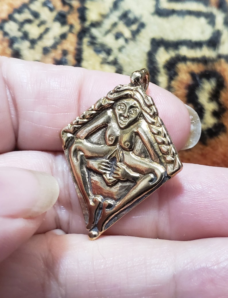 Puede incluir: Un colgante dorado con una escultura en relieve detallada de una figura femenina desnuda con un peinado trenzado. La figura sostiene una flor en su mano y est&aacute; rodeada por un borde trenzado.