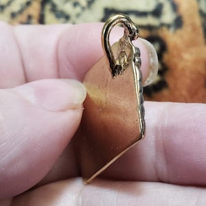 Puede incluir: Un colgante de color dorado con forma cuadrada y un borde decorativo. El colgante tiene un bucle en la parte superior para sujetar una cadena.