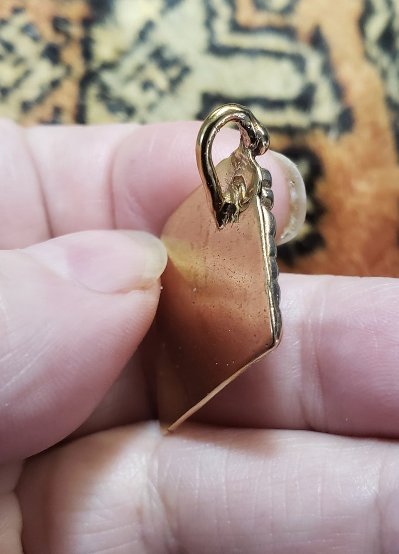 Puede incluir: Un colgante de color dorado con forma cuadrada y un borde decorativo. El colgante tiene un bucle en la parte superior para sujetar una cadena.