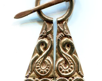 May include: A gold-colored metal brooch with a circular top and two triangular pendants. The pendants are decorated with intricate designs of intertwined snakes. The brooch has a pin that goes through the circle and a loop on the other side.