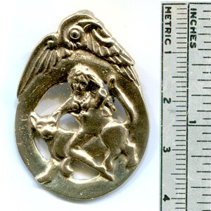 May include: A gold-toned metal pendant featuring a winged figure riding a mythical creature. The pendant is circular and has a detailed design.