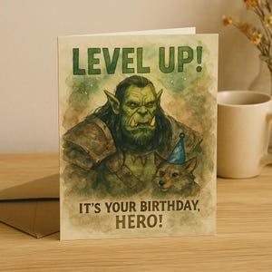 Könnte beinhalten: Geburtstagskarte mit einer Illustration eines Orcs und eines Wolfes mit Partyhut. Der Text "LEVEL UP! IT'S YOUR BIRTHDAY, HERO!" ist grün. Die Karte liegt auf einer Holzoberfläche.