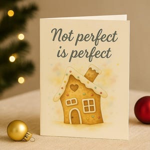 Könnte beinhalten: Eine Grußkarte mit dem Text "Not perfect is perfect" und einer Lebkuchenhaus-Illustration. Eine goldene und eine rote Christbaumkugel liegen auf dem Tisch. Im Hintergrund ist ein Weihnachtsbaum mit Lichtern zu sehen.
