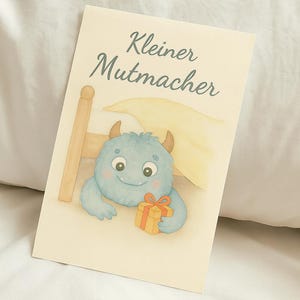 Könnte beinhalten: Eine Grußkarte mit einer Illustration eines blauen Monsters, das ein Geschenk hält. Auf der Karte steht "Kleiner Mutmacher". Der Hintergrund ist cremefarben, mit einem kindlichen Stil.