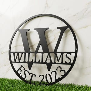 Könnte beinhalten: Schwarzes Metallschild mit Familiennamen, das den Namen "WILLIAMS" und den Buchstaben "W" in einem Kreis zeigt. Das Schild enthält auch "EST 2023". Das Schild wird auf einem Hintergrund aus grünem Gras präsentiert.