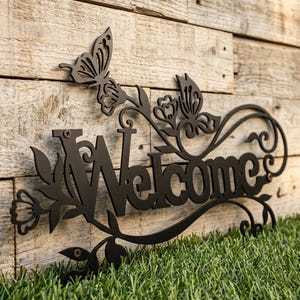 以下が含まれることがあります： 蝶と花の装飾が施された黒い金属製の「Welcome」サイン。サインは木の背景に設置され、緑の草の中に置かれています。「Welcome」の文字は装飾的なフォントです。
