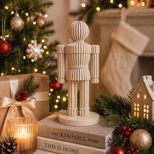 May include: A light beige, slatted wooden nutcracker figurine standing on a circular base. The figure features a rounded head, cylindrical torso, and articulated limbs, all constructed from vertical wooden slats, displayed as a decorative object.
