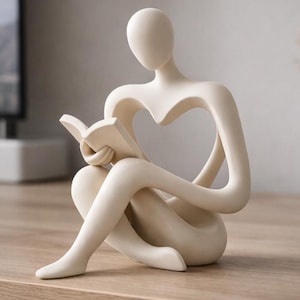 Könnte beinhalten: Eine cremefarbene Skulptur einer sitzenden Person, die ein Buch liest. Die Figur hat einen herzförmigen Ausschnitt im Torso. Die Skulptur steht auf einer Holzoberfläche, mit einem Fernseher und einer Vase im Hintergrund. Die Skulptur ist ein Dekorationsartikel.