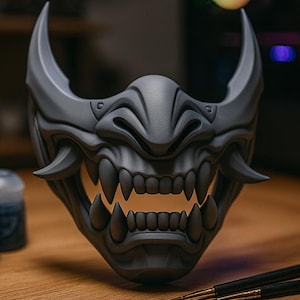 Może przedstawiać: Szara, szczegółowa maska Oni z ostrymi zębami i rogami, spoczywająca na drewnianej powierzchni. W pobliżu znajdują się słoiczki z farbą i pędzle, sugerujące projekt rzemieślniczy lub malarski. Maska jest centralnym punktem, prezentując skomplikowany design.