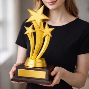 May include: A gold-colored trophy with three rising stars on a dark wooden base. The trophy has a gold plate for engraving. The image shows a woman holding the trophy.