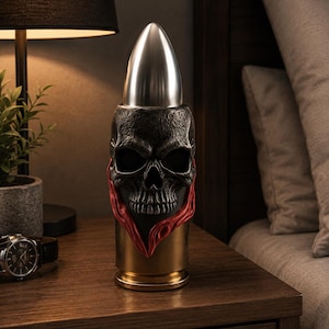 Puede incluir: Objeto decorativo con forma de bala con diseño de calavera. La parte superior es una punta de bala plateada, el centro es una calavera negra con detalles rojos y la base es dorada. Se encuentra sobre una mesita de noche de madera.