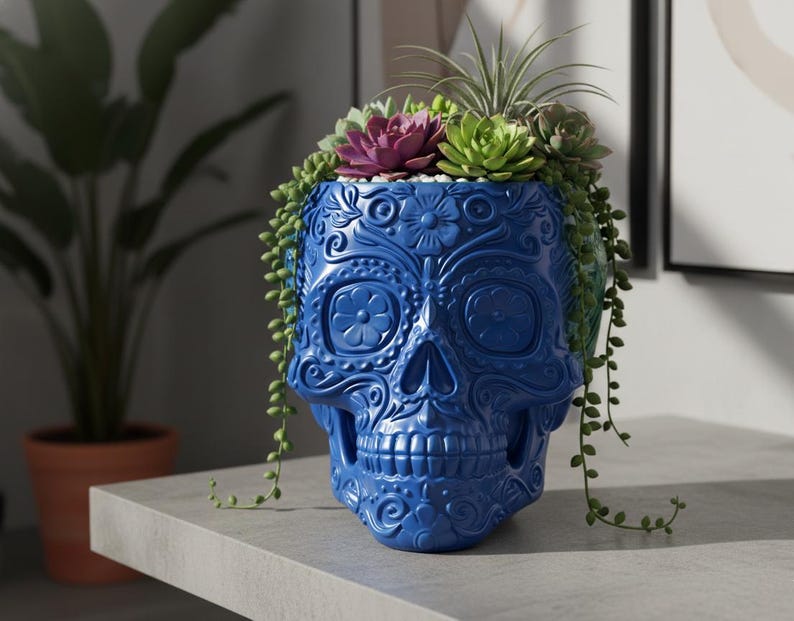 Pu&ograve; includere: Un vaso blu a forma di teschio con intricati disegni floreali, riempito con una variet&agrave; di piante grasse e piante ricadenti. Il vaso &egrave; in ceramica o materiale simile e si trova su una superficie grigia.
