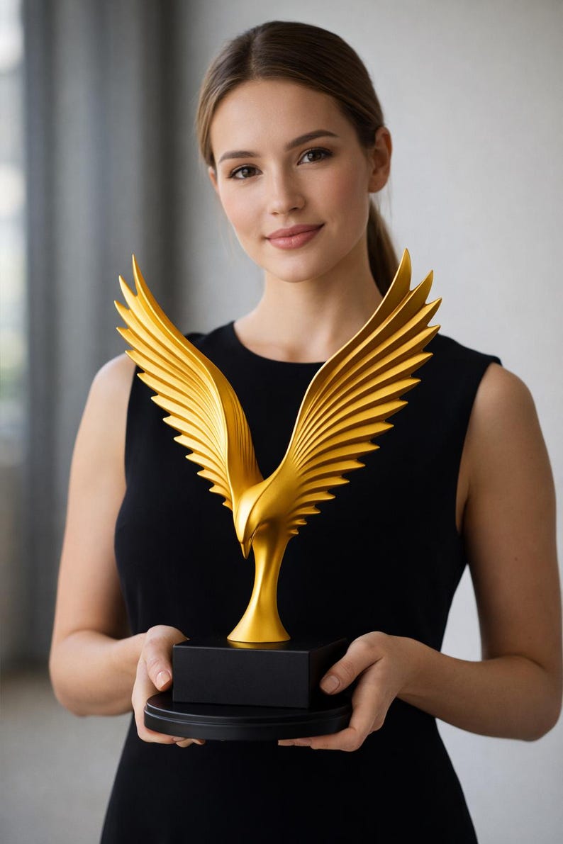 May include: A gold-colored eagle sculpture with outstretched wings, mounted on a black rectangular base and a round base. The sculpture has detailed feathering and is held in front of a person wearing a black dress.