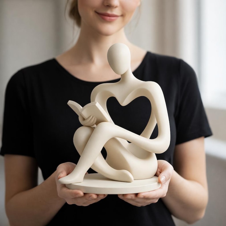 May include: A cream-colored sculpture of a person reading a book. The figure is seated with legs crossed and arms holding the book. The body has a heart-shaped cutout. The sculpture is on a round base.