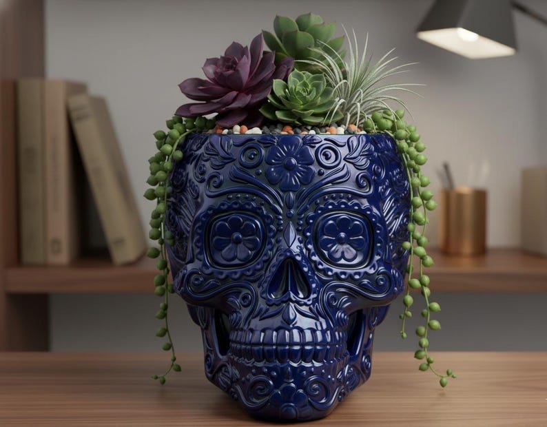 Pu&ograve; includere: Un vaso di ceramica blu scuro a forma di teschio con intricati disegni floreali. Il vaso &egrave; riempito con una variet&agrave; di piante grasse, tra cui una succulenta viola scuro, una succulenta verde e piante verdi pendenti. Il vaso &egrave; su una superficie di legno.
