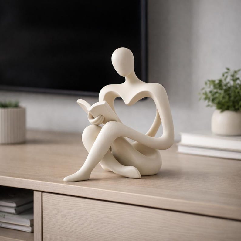 May include: A cream-colored sculpture of a person reading a book. The figure is seated with legs crossed, holding the book open. The design features a heart-shaped cutout in the torso. The sculpture is placed on a light-colored wooden surface.