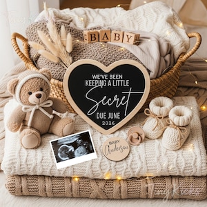 Puede incluir: Una pizarra en forma de corazón con el texto "WE'VE BEEN KEEPING A LITTLE SECRET DUE JUNE 2026" está rodeada de artículos para bebés. Un osito de peluche, una imagen de ultrasonido, patucos y un chupete también están presentes. Un letrero de madera dice "Baby Anderson".