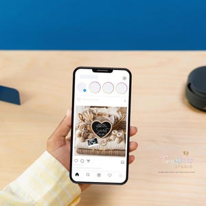 May include: A smartphone displays a digital pregnancy announcement. The announcement features a heart-shaped chalkboard with the words "We've been keeping a little secret" and "Due June". The image includes a teddy bear, baby shoes, and a sonogram.