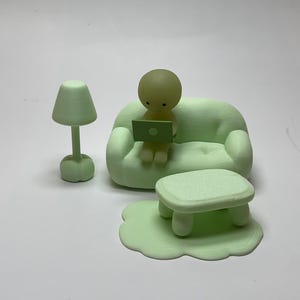 May include: A light green miniature living room set with a sofa, coffee table, rug, lamp, and a figure using a laptop. The set is made of a solid material and has a smooth finish. The figure is sitting on the sofa.