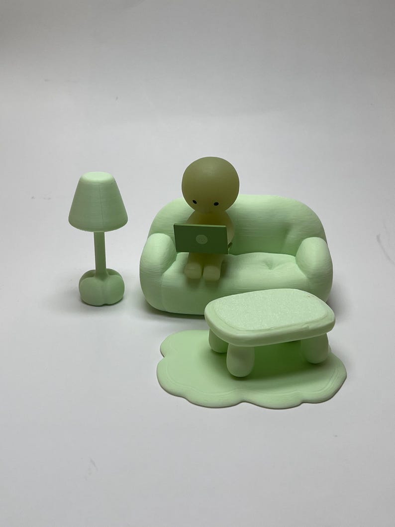 May include: A light green miniature living room set with a sofa, coffee table, rug, lamp, and a figure using a laptop. The set is made of a solid material and has a smooth finish. The figure is sitting on the sofa.