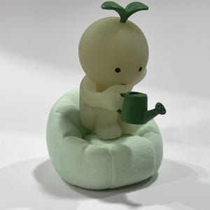 Puede incluir: Figura verde claro de un personaje peque&ntilde;o y redondo con un brote en la cabeza, sentado en un coj&iacute;n texturizado verde claro. El personaje sostiene una peque&ntilde;a regadera verde, lo que sugiere un tema de jardiner&iacute;a.