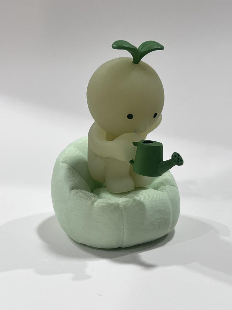 Puede incluir: Figura verde claro de un personaje peque&ntilde;o y redondo con un brote en la cabeza, sentado en un coj&iacute;n texturizado verde claro. El personaje sostiene una peque&ntilde;a regadera verde, lo que sugiere un tema de jardiner&iacute;a.