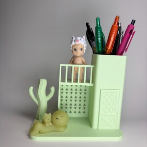 May include: A light green desk organizer with a pen holder, a small balcony, and a cactus-shaped accent. A small figurine wearing a sheep hat stands on the balcony. A figurine of a person and a cat are lying down. Assorted pens are in the pen holder.