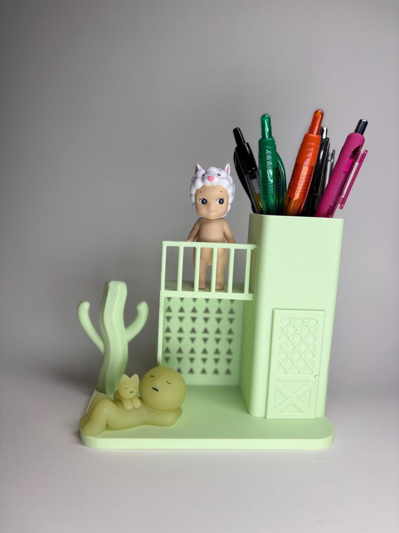 May include: A light green desk organizer with a pen holder, a small balcony, and a cactus-shaped accent. A small figurine wearing a sheep hat stands on the balcony. A figurine of a person and a cat are lying down. Assorted pens are in the pen holder.