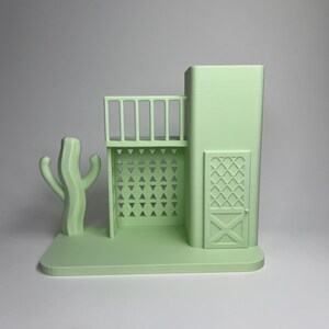 May include: A light green 3D-printed model of a Southwestern-style building with a balcony, a cactus, and a door with an X-shaped design. The model is set on a rectangular base and has a minimalist aesthetic.