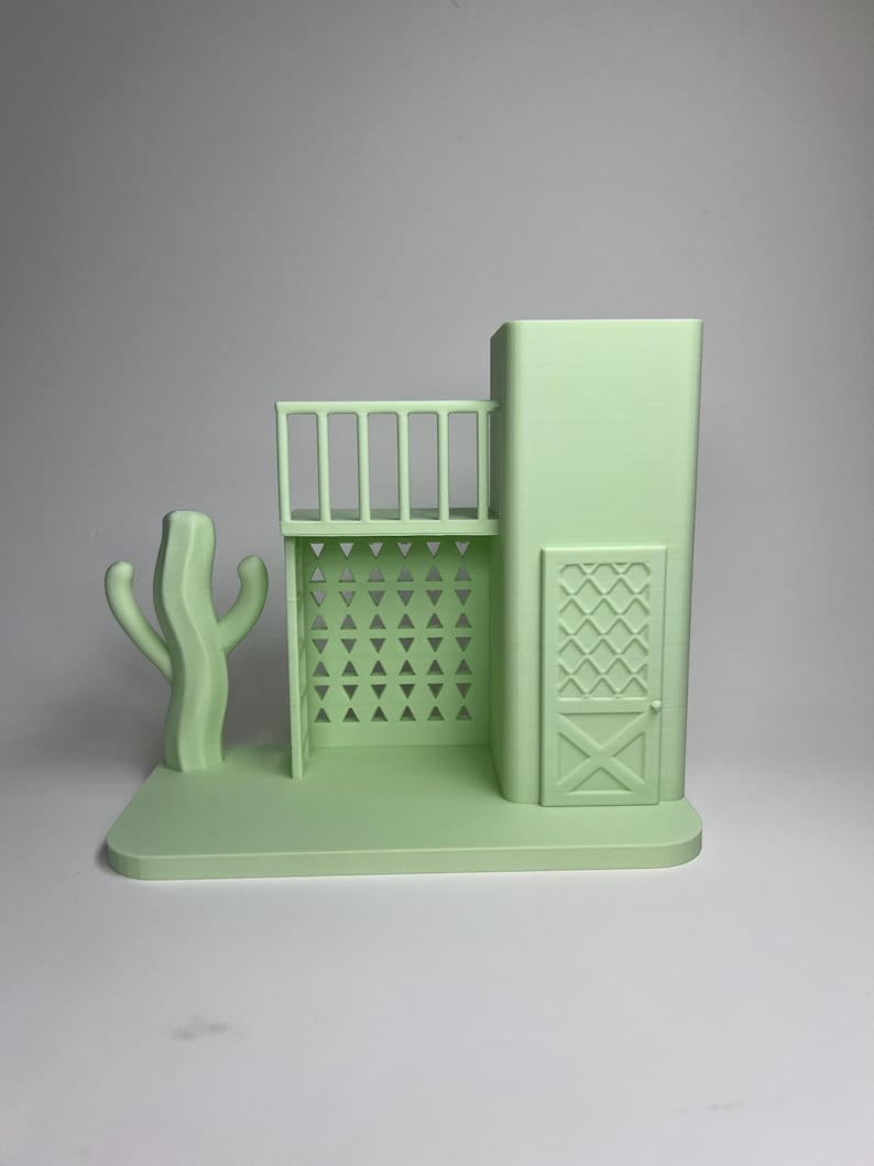 May include: A light green 3D-printed model of a Southwestern-style building with a balcony, a cactus, and a door with an X-shaped design. The model is set on a rectangular base and has a minimalist aesthetic.