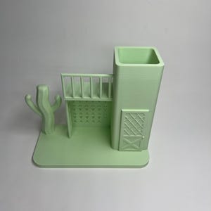 May include: A light green 3D-printed desk organizer shaped like a Southwestern-style building with a cactus, balcony, and door. The organizer has a rectangular base and a tall, square container for pens or pencils.