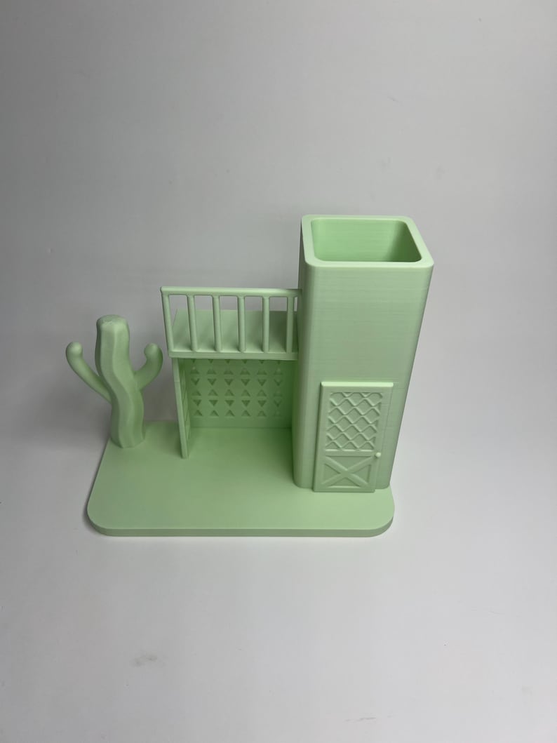 May include: A light green 3D-printed desk organizer shaped like a Southwestern-style building with a cactus, balcony, and door. The organizer has a rectangular base and a tall, square container for pens or pencils.
