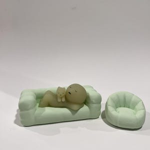 Puede incluir: Un sof&aacute; en miniatura verde claro con una figura y una figura de gato. La figura est&aacute; acostada en el sof&aacute;, con una figura de gato descansando sobre su pecho. Al lado del sof&aacute; hay un sill&oacute;n en miniatura verde claro a juego.