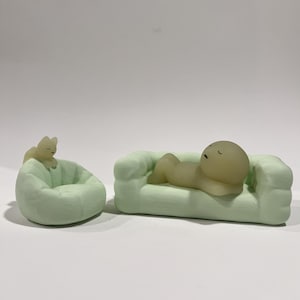 Puede incluir: Un sof&aacute; en miniatura verde claro con una figura acostada sobre &eacute;l, y un puf verde claro con una peque&ntilde;a figura sentada sobre &eacute;l. Las figuras son de color amarillo claro. El fondo es blanco.