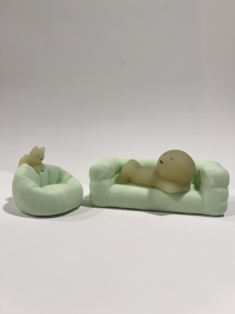 Puede incluir: Un sof&aacute; en miniatura verde claro con una figura acostada sobre &eacute;l, y un puf verde claro con una peque&ntilde;a figura sentada sobre &eacute;l. Las figuras son de color amarillo claro. El fondo es blanco.