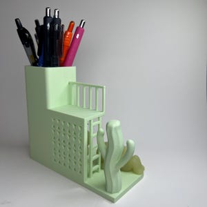 May include: A light green desk organizer with a tiered design, featuring a small balcony and a cactus sculpture. The organizer holds various pens and pencils. The design includes a ladder and geometric cutouts.